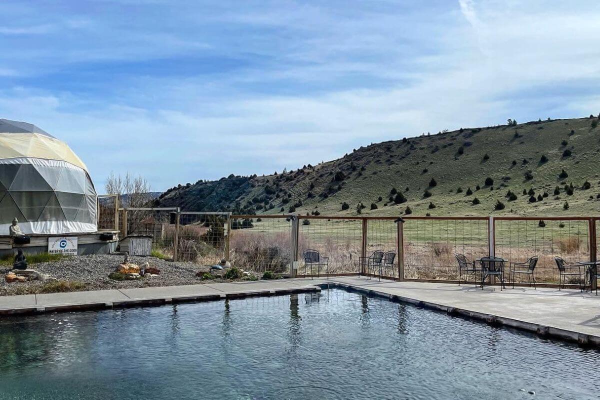The Norris Hot Springs pool, enclosed by a fence, offers a view of distant grassy hillsides.