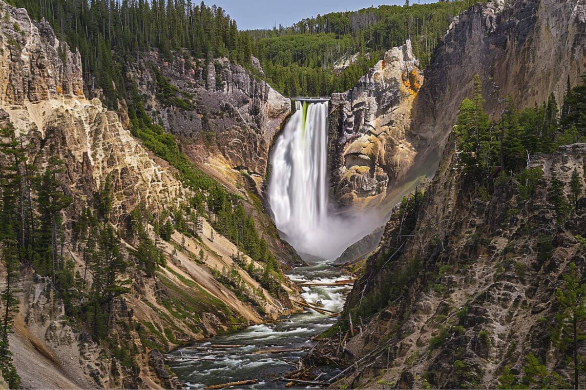 Lower Falls cascades down a cliff into a river that cuts through a steep, forested canyon in Montana.
