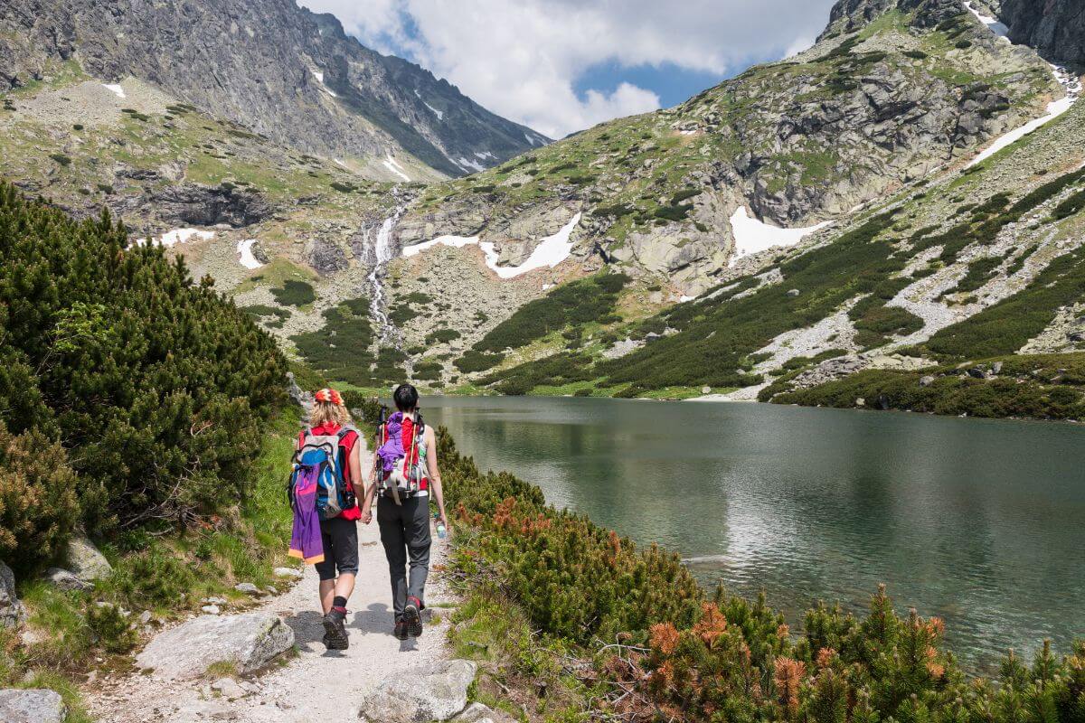 Hikers trek alongside a scenic mountain trail, passing a tranquil alpine lake with snow remnants on the slopes in the background.