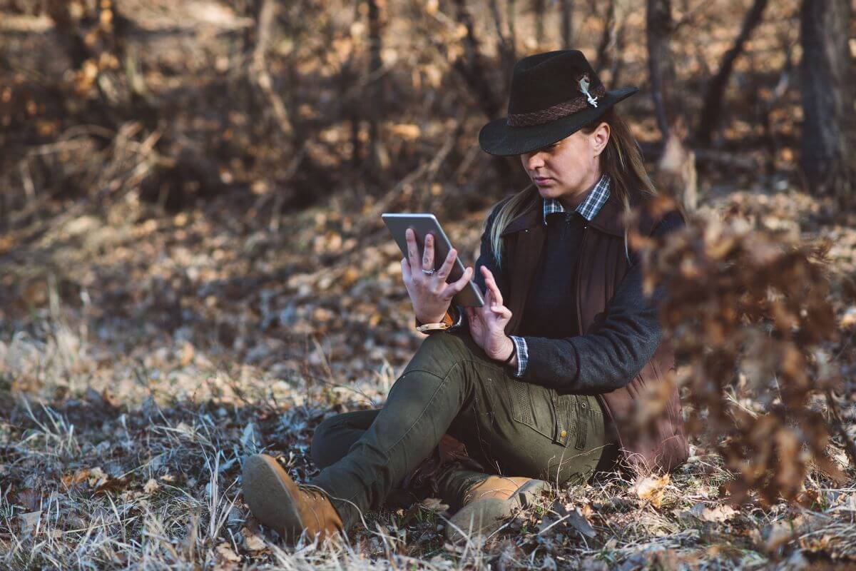 A woman wearing a hat sits on the ground in a forest, reading Montana hunting regulations on her device.
