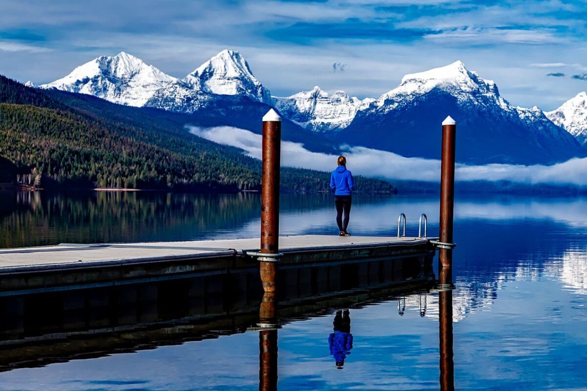 A solo traveler in Montana stands at the edge of a dock, admiring the view of the lake and mountains.