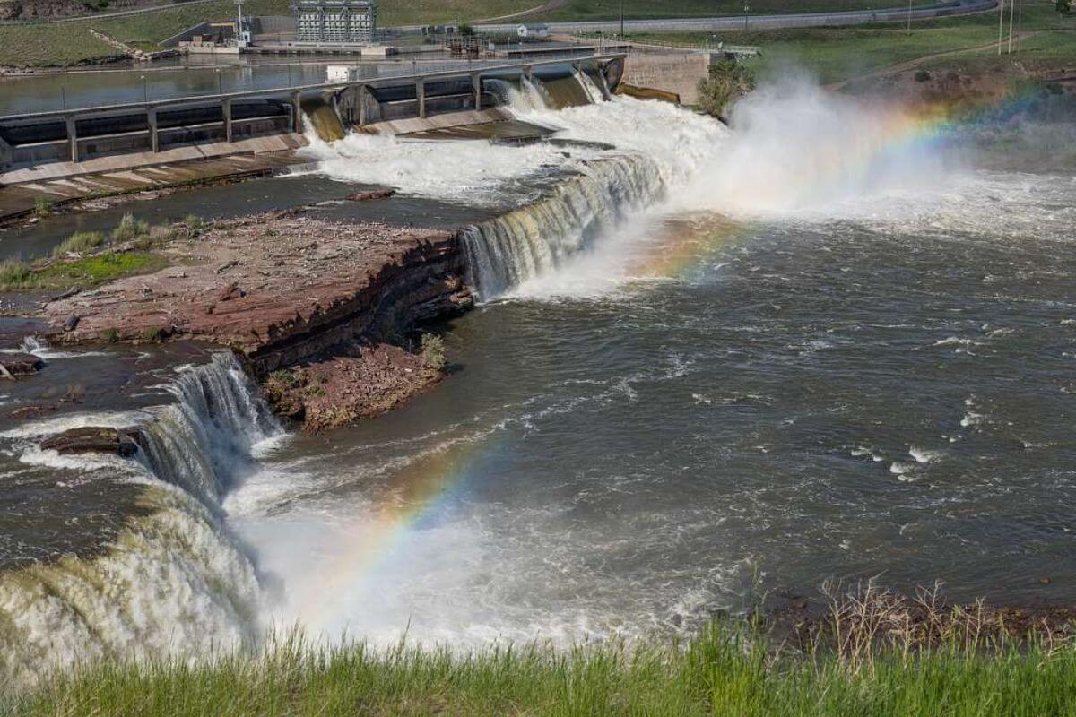 Rainbow Falls near a dam in Montana flows over rocks, with a small rainbow visible in the mist under a clear sky.