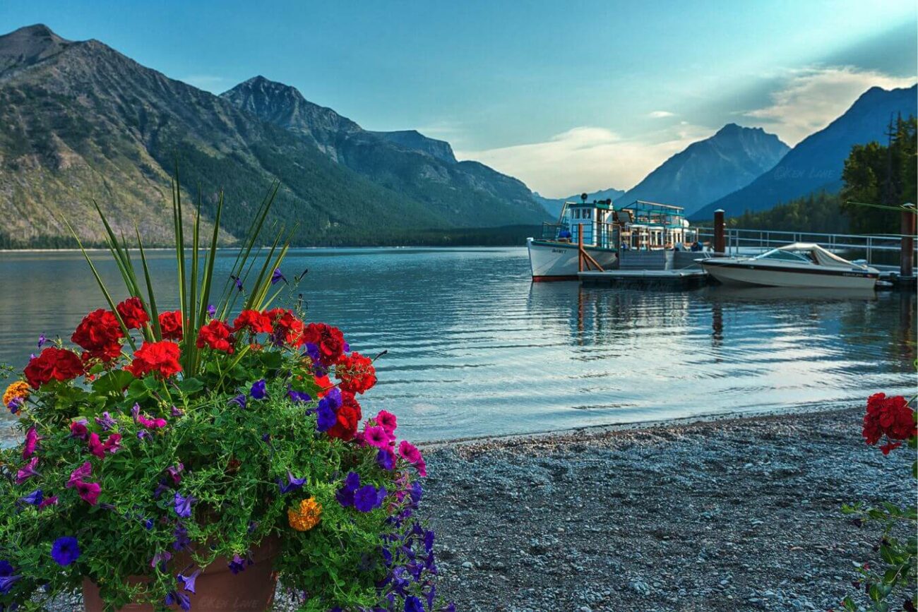 A scenic view of a quiet lake in Montana, bordered by mountains, featuring a small dock with a boat and vibrant flowers