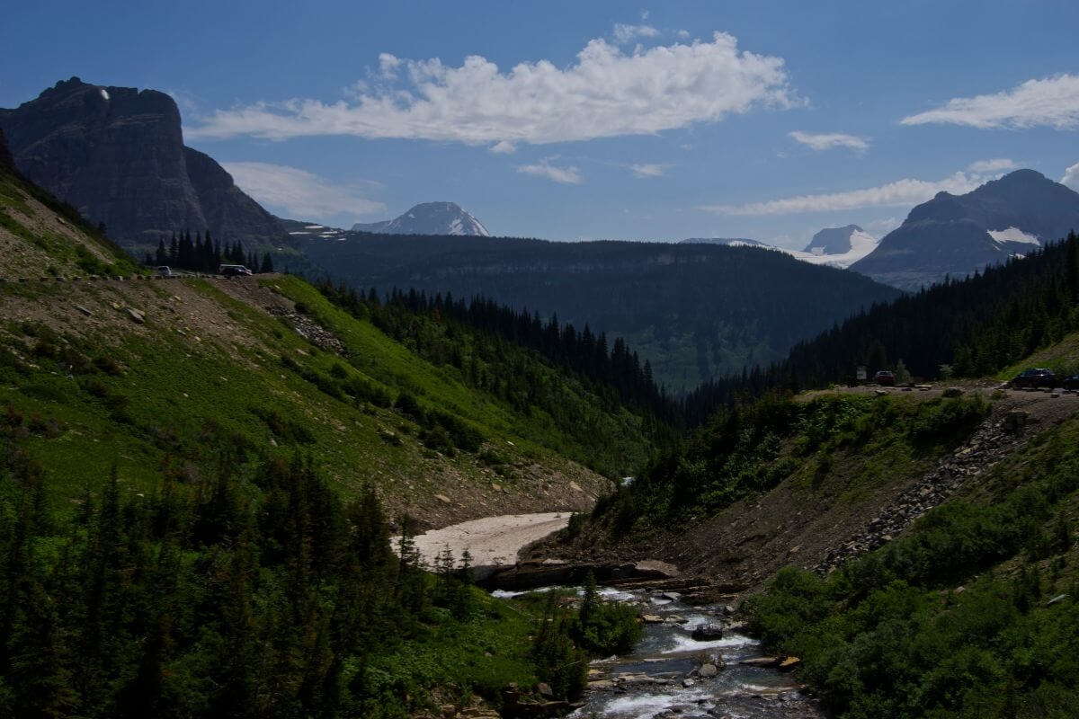 A river runs through a lush area of a mountain near a road, with scenic peaks in the background.