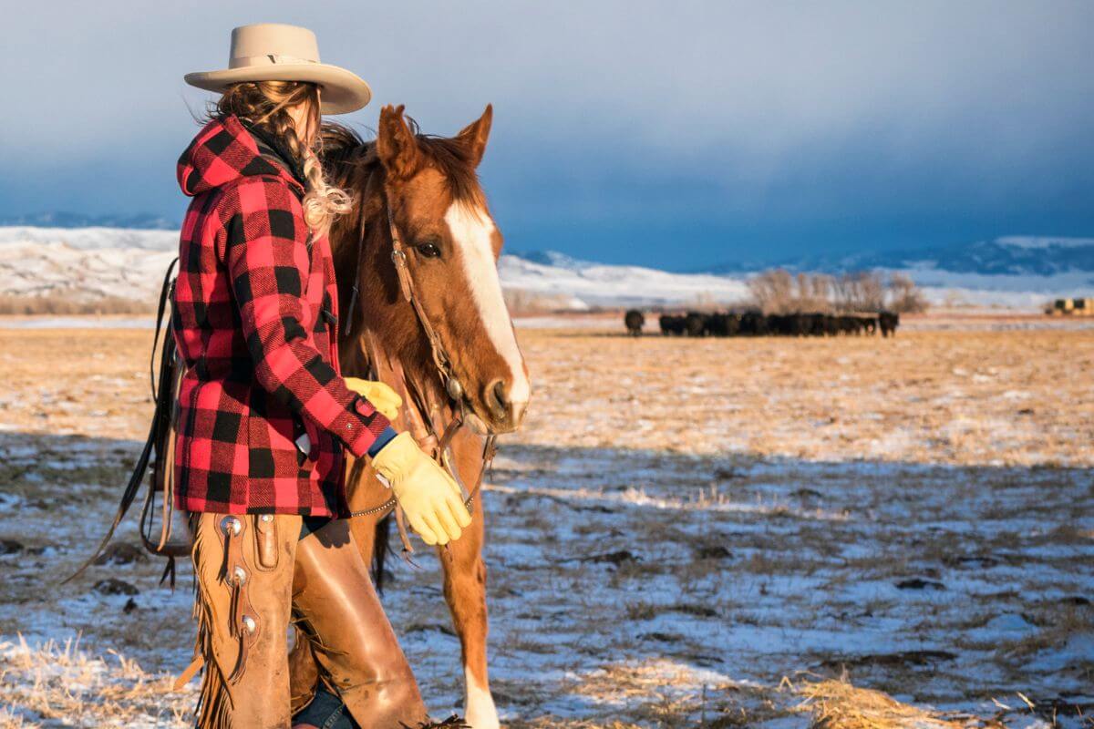 A woman in a cowboy hat standing next to a horse in a snowy field on a Montana ranch.