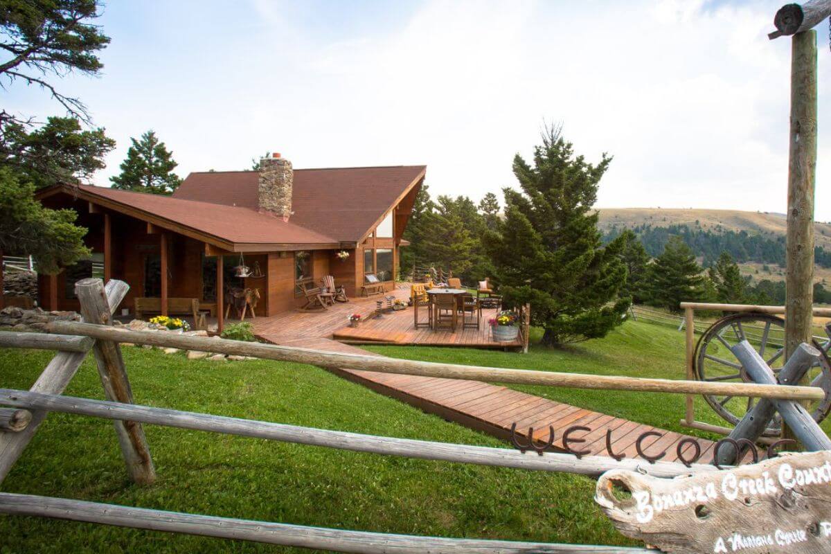 A rustic cabin at Bonanza Creek Country Guest Ranch has a wooden deck with outdoor seating and vibrant flowers. It is surrounded by lush greenery and tall trees. A wooden fence with a partially visible "Welcome" sign frames the foreground.