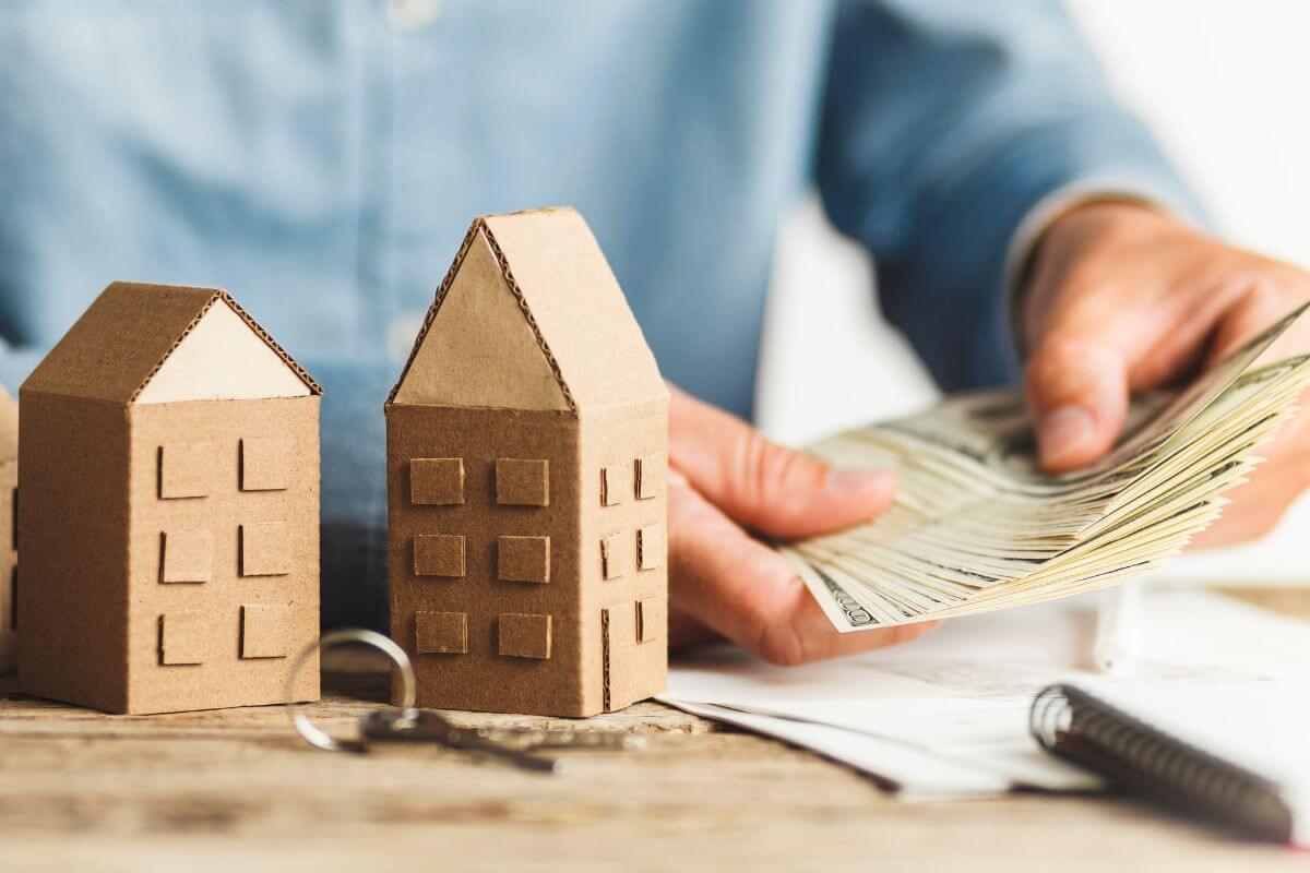 A man holding money with a cardboard model of a house and keys beside it