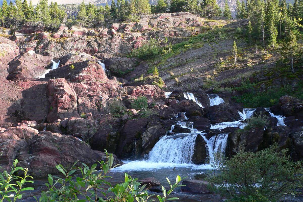 Red Rock Falls cascades through large red rocks in a lush wooded area in Glacier National Park.