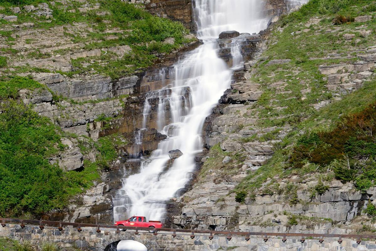 A red pickup truck drives past the majestic Bird Woman Falls along Going-to-the-Sun Road.