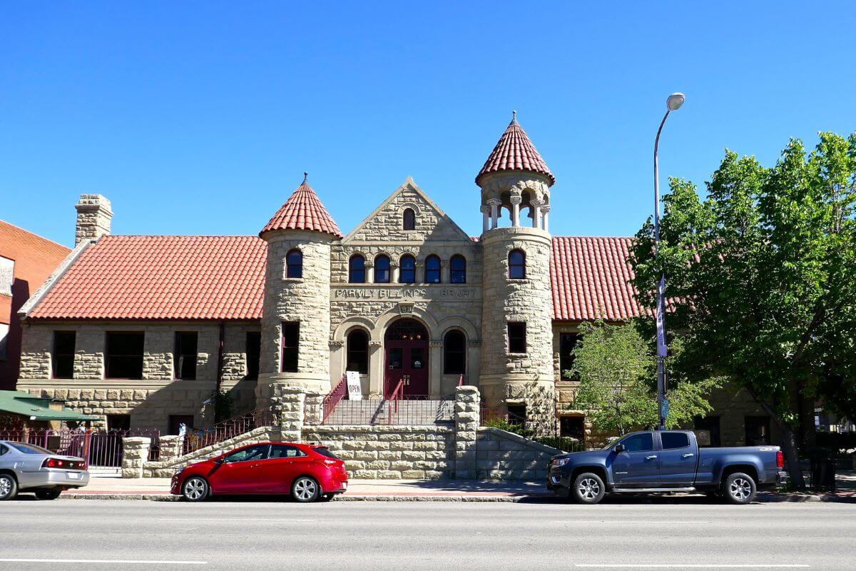 A large stone building in Montana, visited by tourists with cars parked in front of it.
