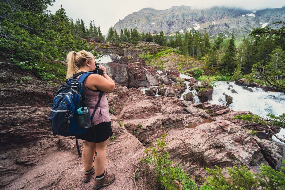 Woman with a backpack photographs a creek and mountains in a forested area in a trail going to Redrock Falls.