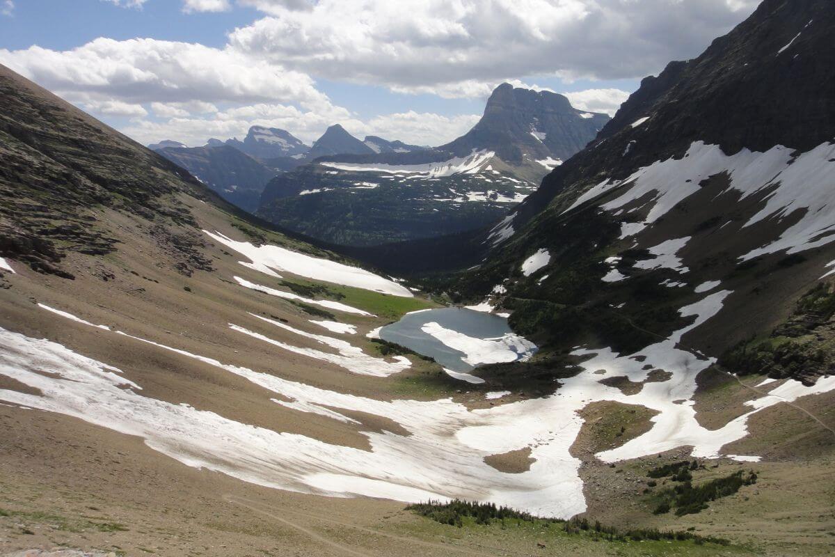 A scenic mountain landscape in Montana near Ptarmigan Waterfall with snow patches, a small blue lake, distant peaks, and rolling clouds under a vast sky.