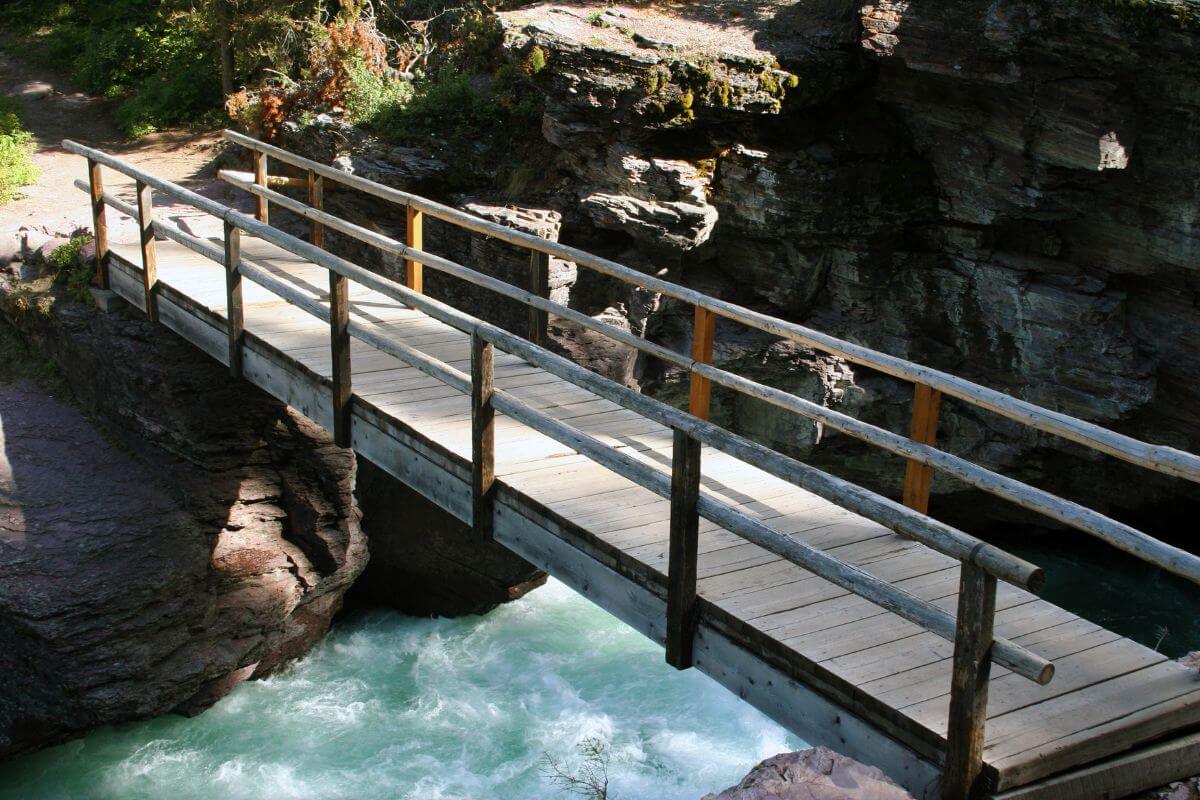 A wooden bridge with railings spans a flowing river in a rocky, forested area near Saint Mary Waterfall, Montana. 