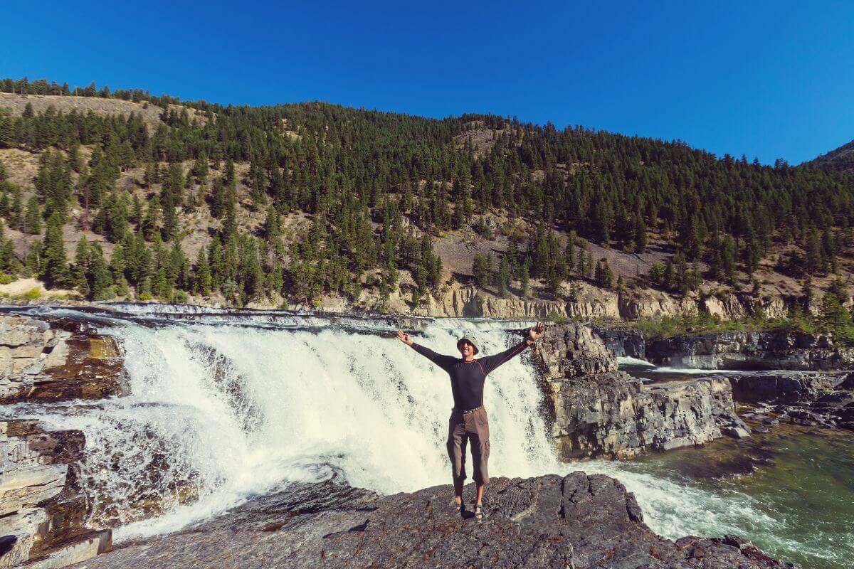 A tourist poses for a picture on a nearby cliff, with the cascading waters of Kootenai Falls in the background.