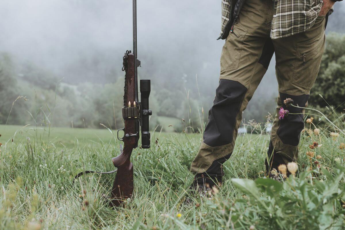 A person stands in a grassy field with a rifle propped up next to them, getting ready to buy Montana preference points.