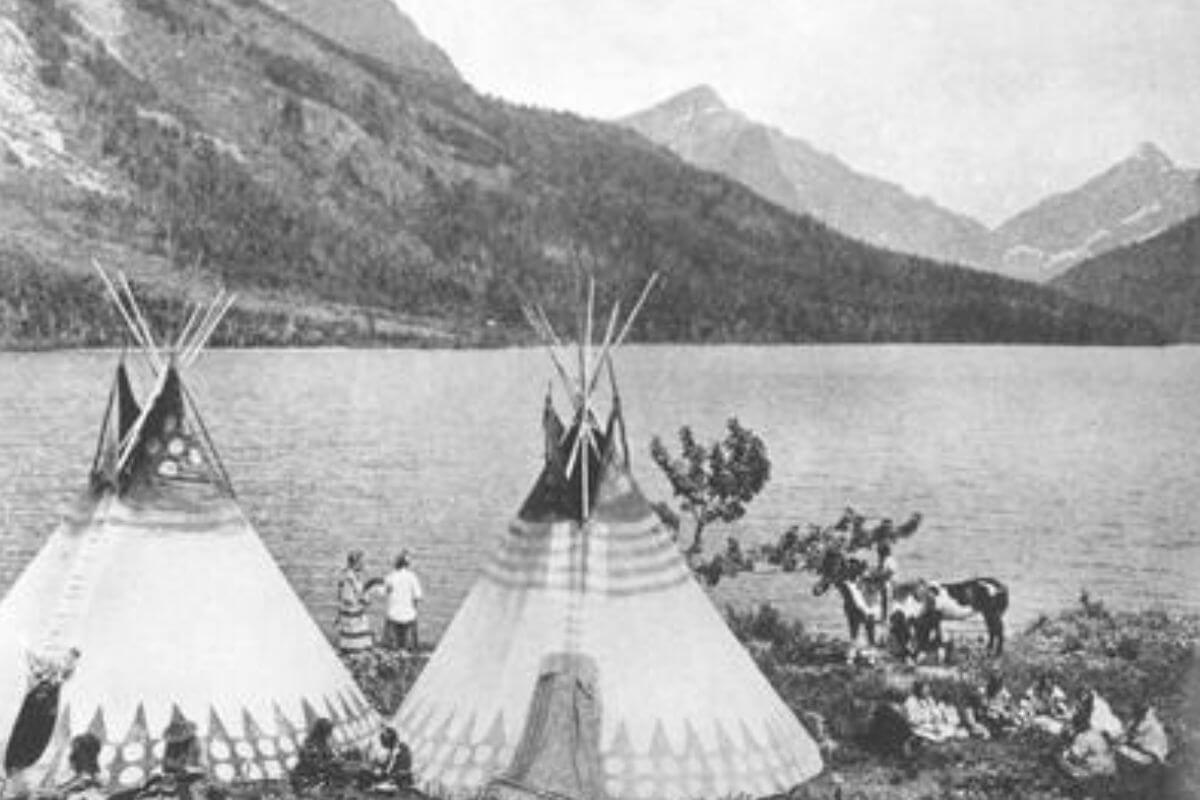 A black and white image of teepees in Montana in front of a lake with mountains in the background.