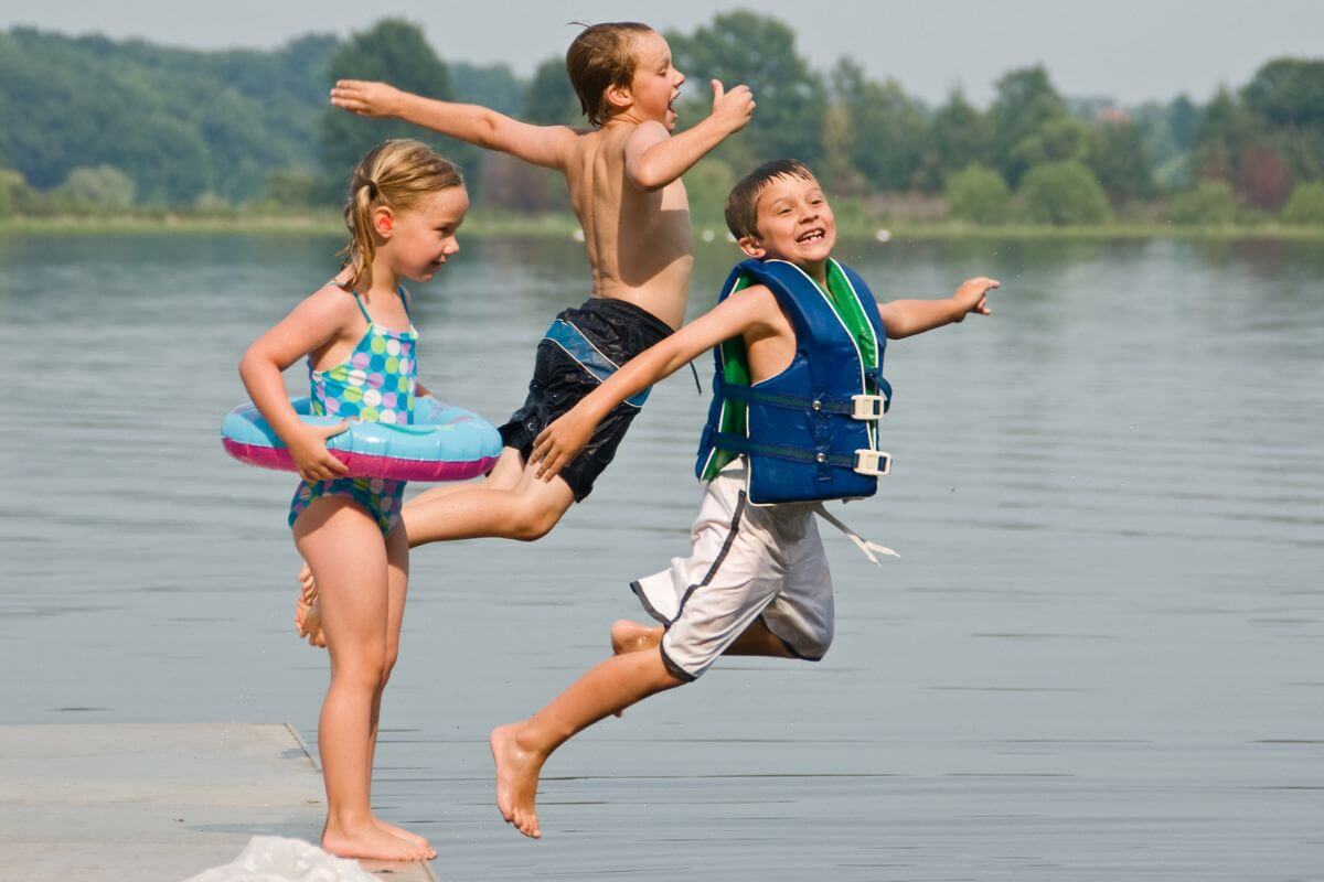 Three children in swimwear gleefully jumping into a lake near Martin Waterfall in Montana, on a sunny day, embracing the joys of summer.