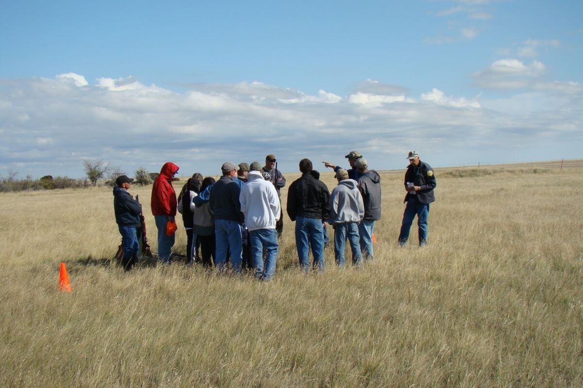 A group of people wearing casual outdoor clothing are standing in a circle in a grassy field at Benton Lake Wildlife Refuge in Montana.