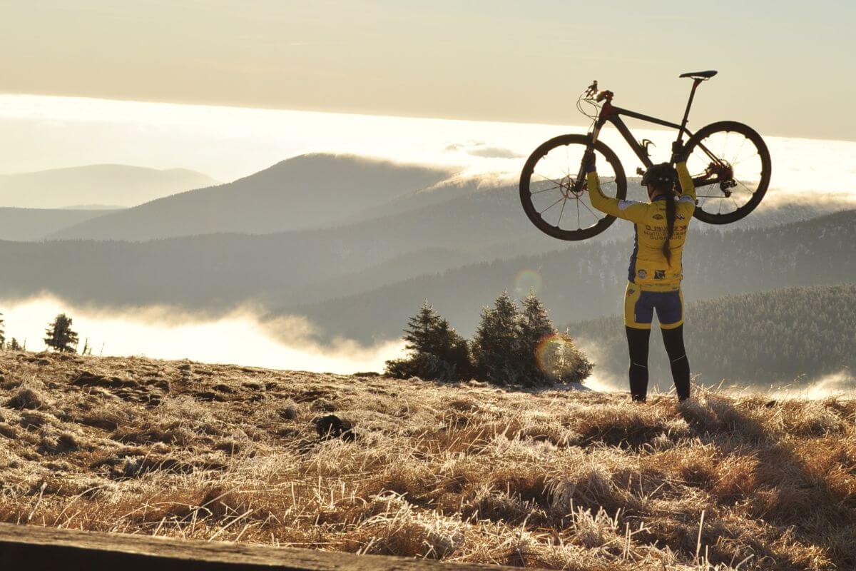 A cyclist standing atop a frosty mountain in Montana holds their bike overhead against a backdrop of cloud-covered hills illuminated by the soft light of sunrise.