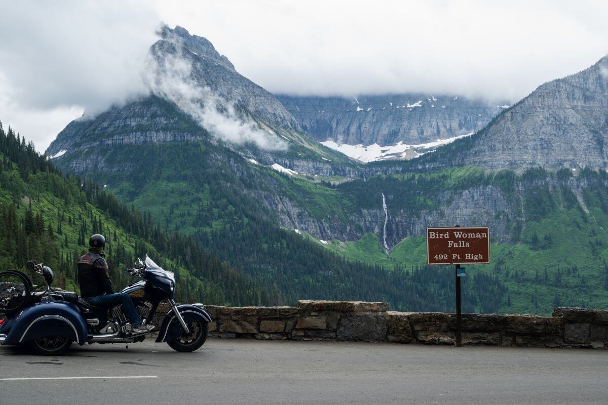 A motorcyclist beside a sign for "Bird Woman Falls 492 ft." at Glacier National Park, perfect for Montana motorcycle tours.