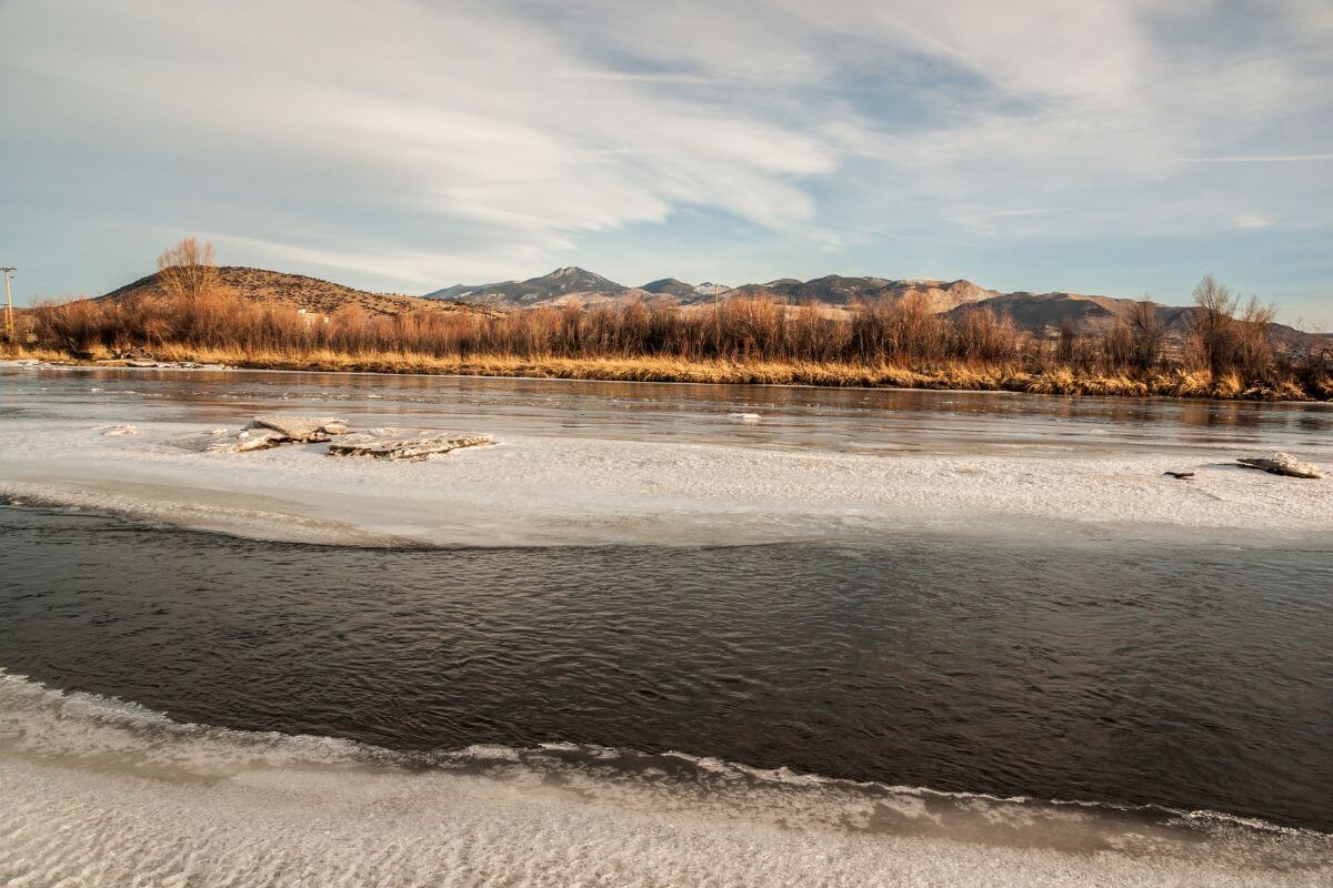 A frozen river framed by bare trees near Jefferson Creek Trailhead, with Montana's majestic mountains in the distance under a golden sky.