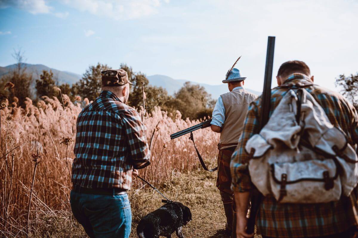 Three men walk through a field with a black dog, carrying hunting gear and rifles, and discuss applying for Montana preference points as a group.