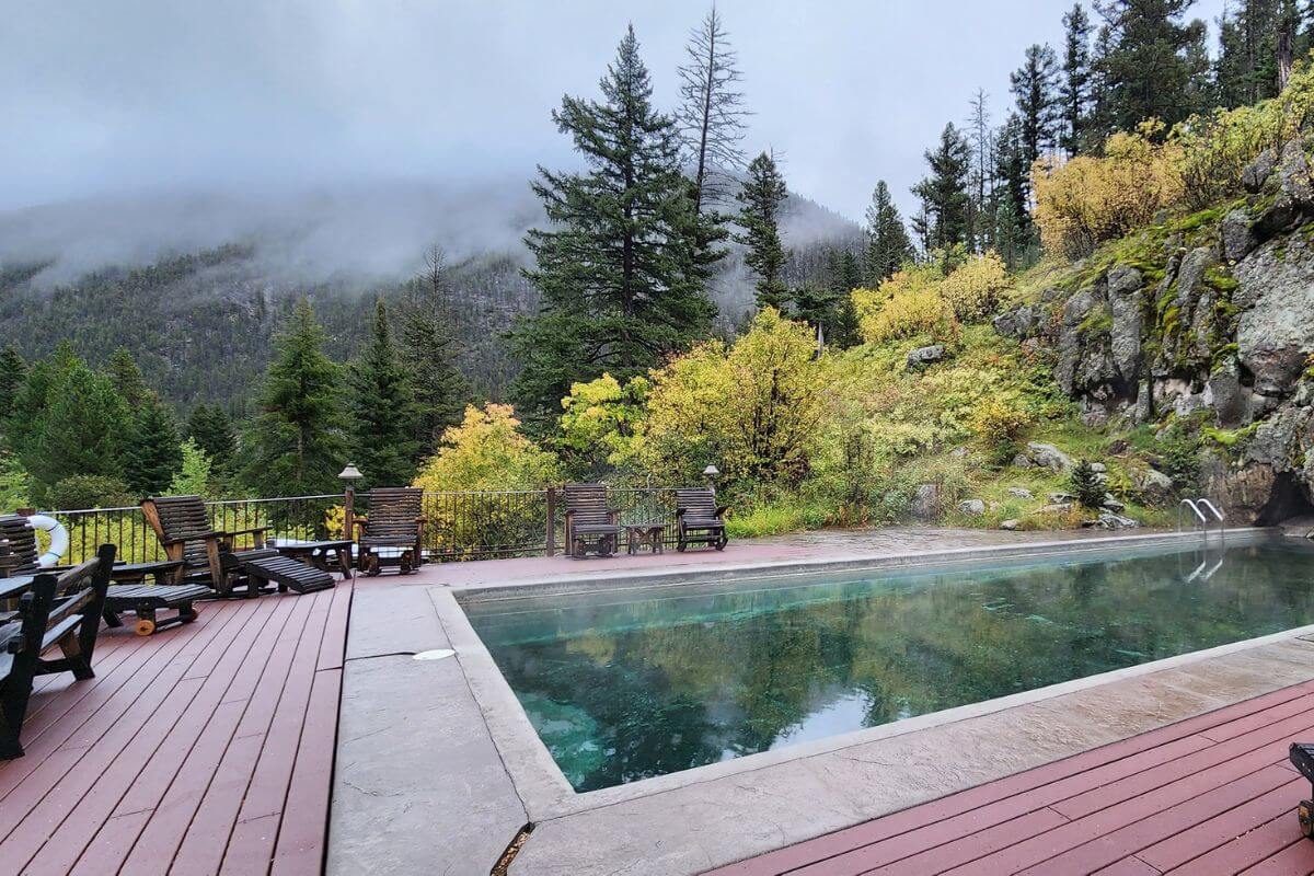 A Potosi Hot Springs pool, surrounded by a steel fence and complemented with wooden lounge chairs, sits amid the Montana wilderness.