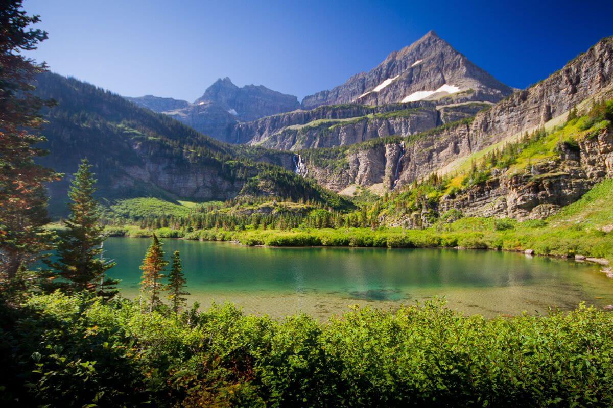 Stoney Indian Pass Trail with a serene mountain lake with clear water surrounded by lush green foliage and towering peaks under a clear blue sky.