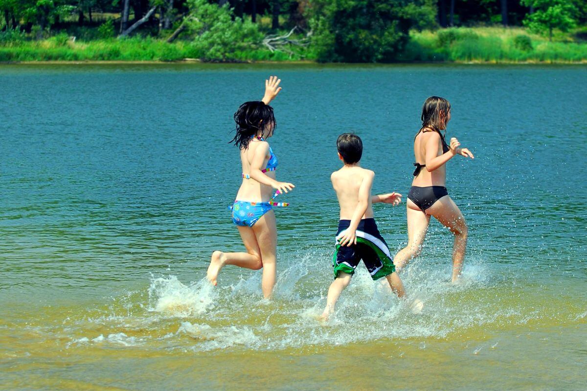 Three children running into a lake, splashing water around, on a bright sunny day with trees.