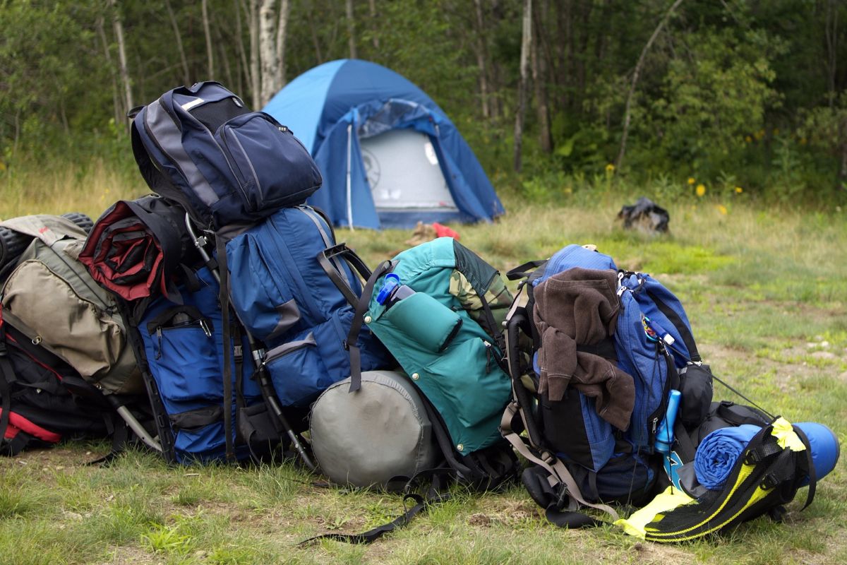 A collection of backpacks with camping gear piled up in a grassy area, with a blue dome tent in the background surrounded by trees near Mill Falls, Montana.