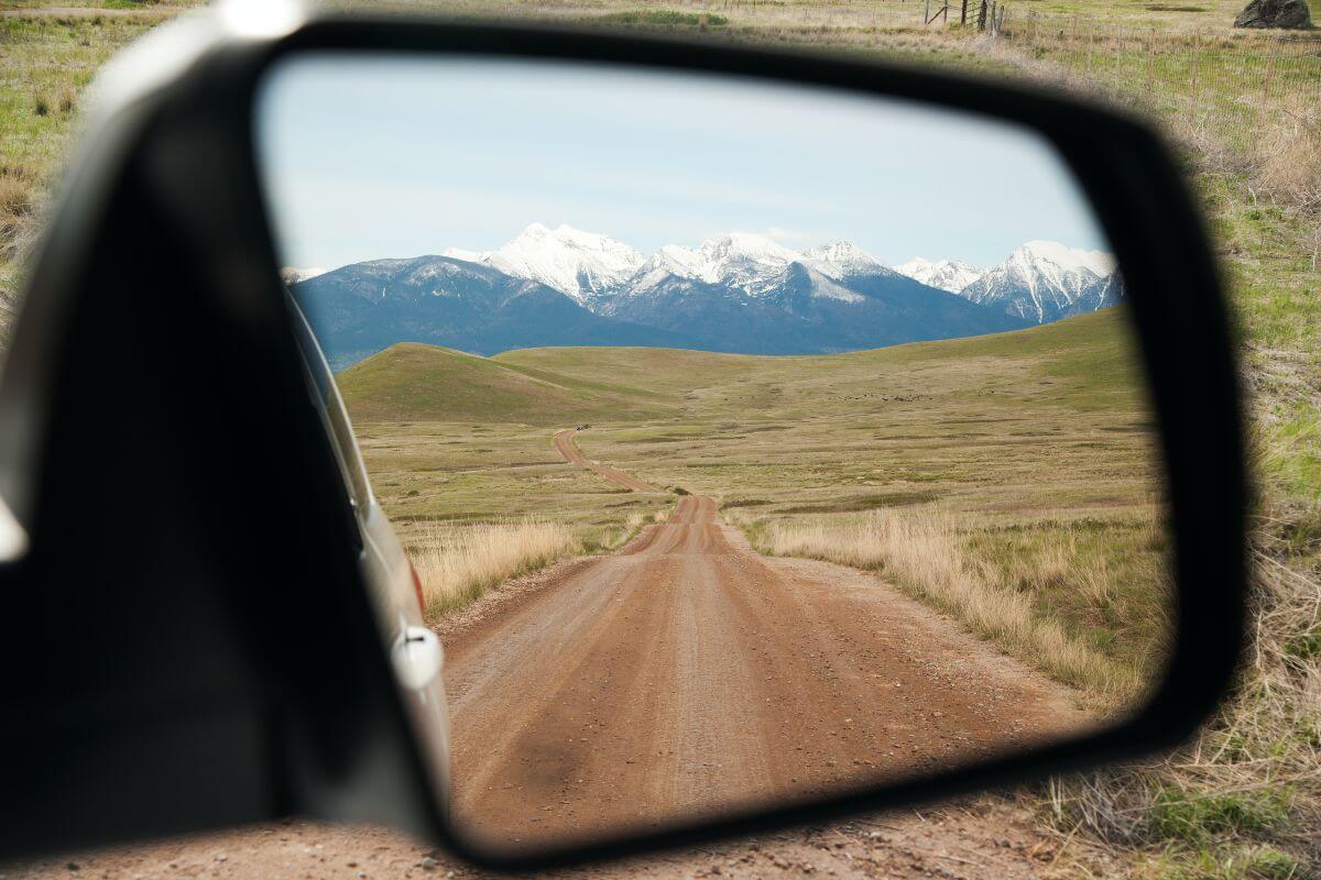 View of a dirt road at the Bison Range Wildlife Refuge in Montana, leading into a valley surrounded by snow-capped mountains as seen through a car's side mirror.