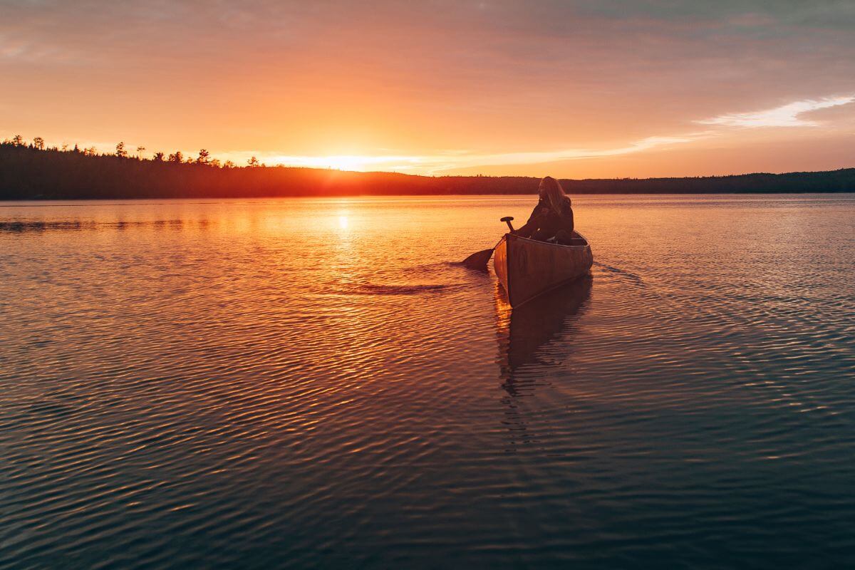 A person in a canoe paddles on a calm lake during a stunning sunset, with orange and red hues reflected in the water and silhouetted trees.