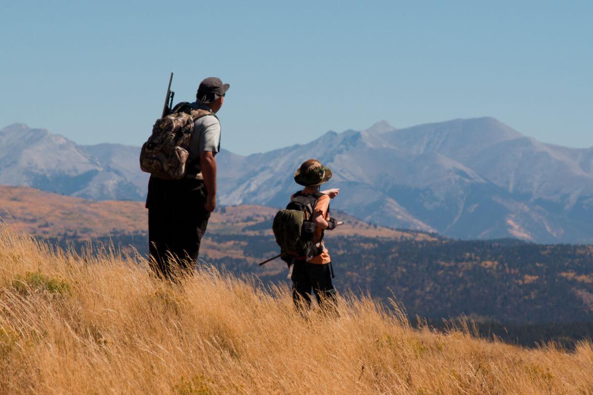 Two hikers with backpacks stand in a grassy field and looking at distant mountain ranges, getting ready for Montana elk hunting.