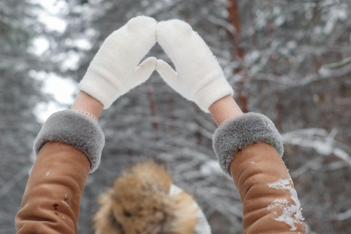 A woman with winter gloves is holding her hands up in the air in a snowy forest.