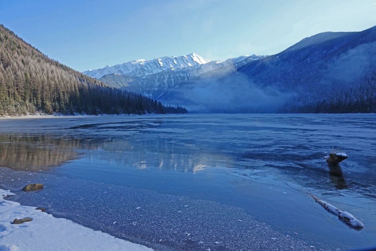 A serene winter landscape featuring a partially frozen lake with fog above it, surrounded by snow-covered pine trees and Stanton Mountain in the distance 