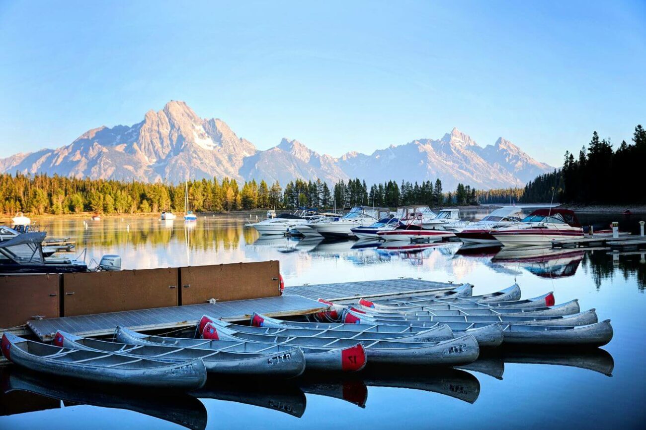 A peaceful twilight scene with canoes and boats docked on a calm lake in Montana, reflecting clear skies and the rugged peaks of the Teton range in the background.