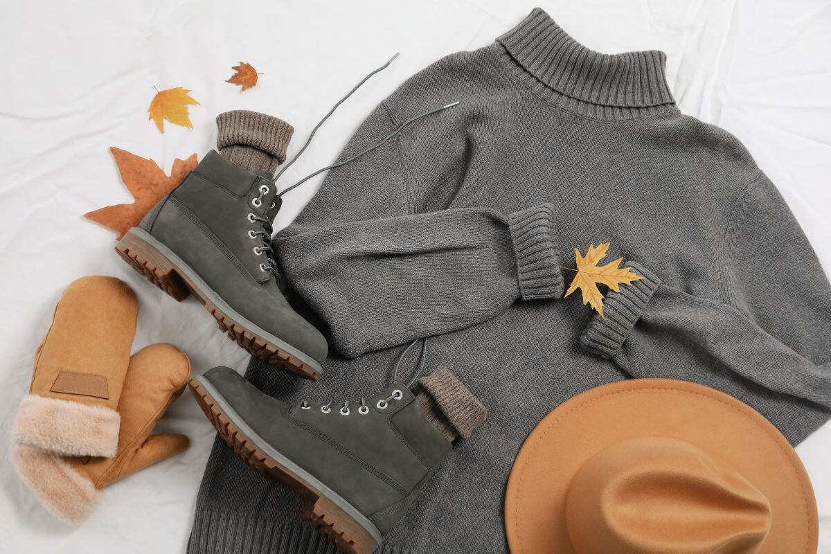 Fall clothing essentials, including sweaters and boots, are laid out on a bed alongside some fall leaves.