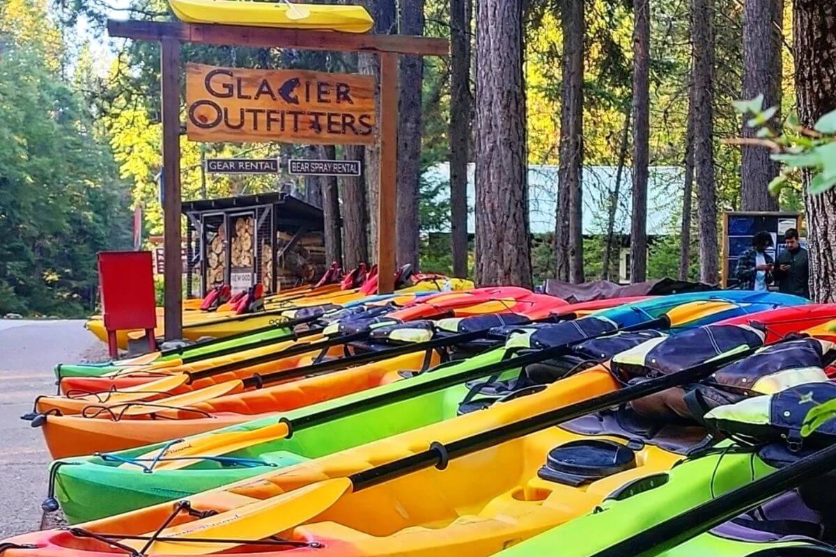 Colorful kayaks are lined up near the glacier outfitters store in a wooded area in Montana, with a gear rental sign visible above them