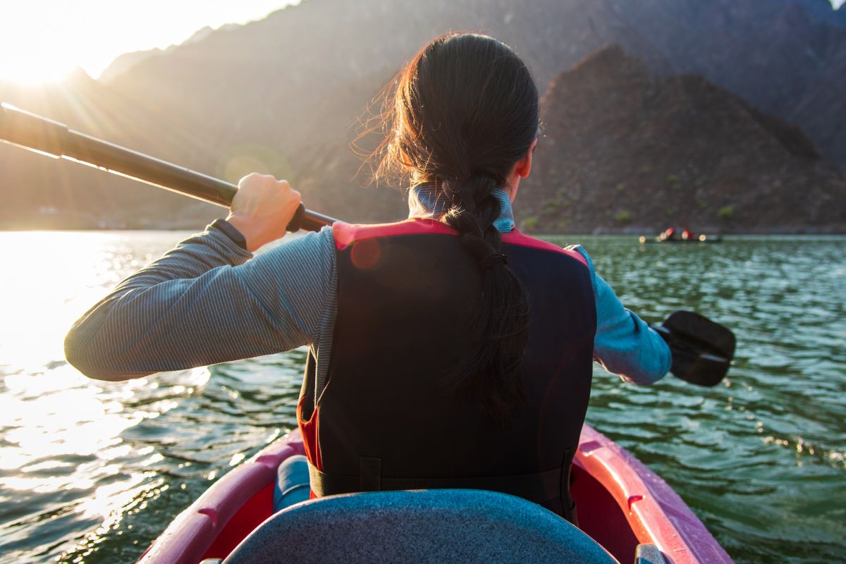 A woman in a life vest kayaks on a lake with mountains in the background