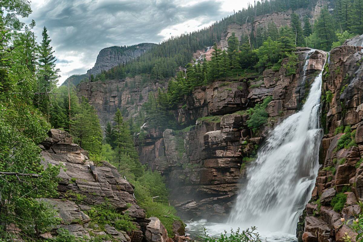 The Mission Falls cascades down rugged cliffs amid a lush, green forest under a partly cloudy sky in Montana.