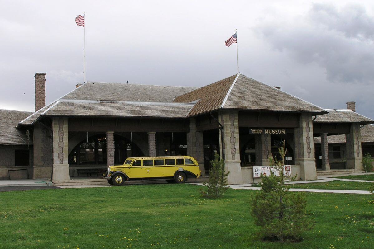 A yellow bus parked in front of a stone museum building with two American flags under an overcast sky in Montana.