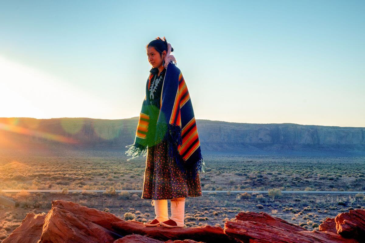 A Native American woman stands on a rocky path in a reservation.