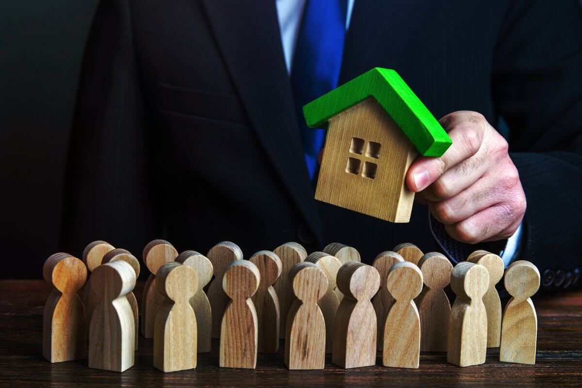 A businessman is holding a house model in front of a group of wooden figures