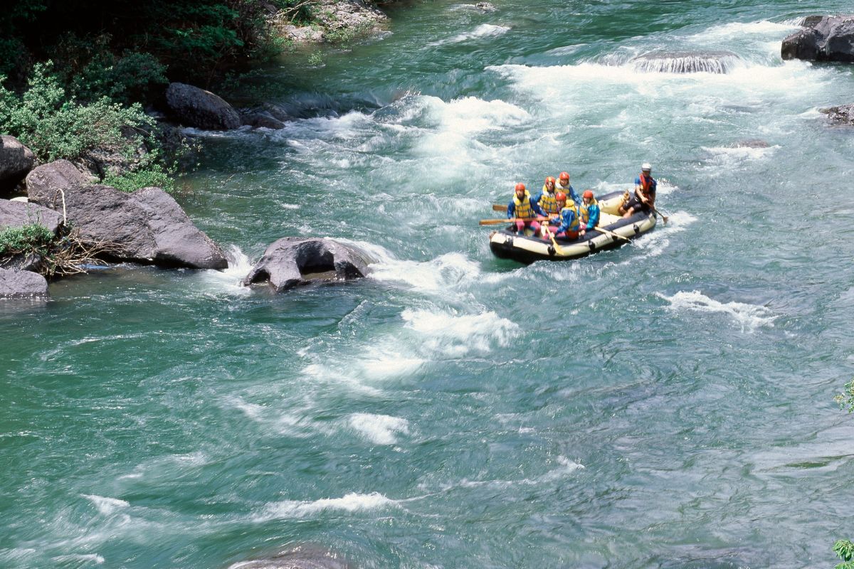 A group in helmets and life jackets rafts on a rocky river near Pintler Waterfall, Montana, surrounded by greenery.