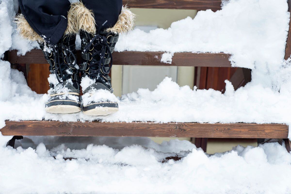 A person wearing winter boots with fur stands on snowy outdoor steps