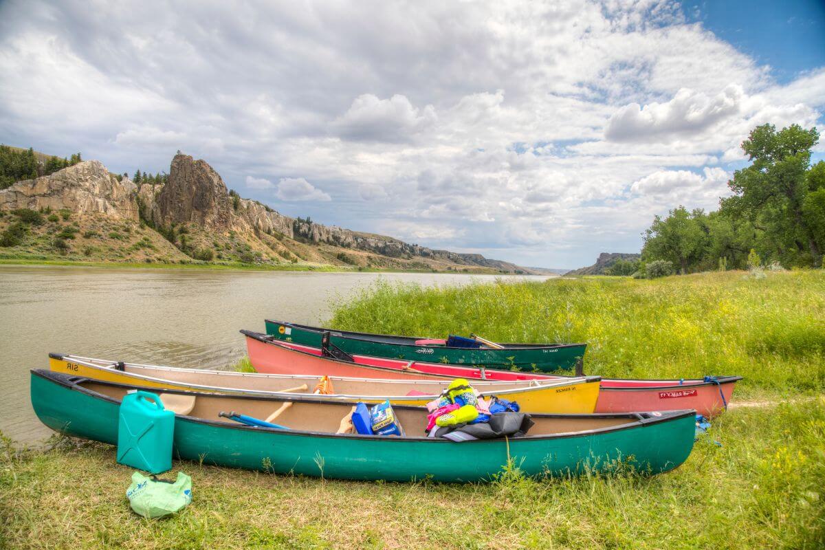 Four differently colored canoes with supplies are lined up by the Missouri River bank, ready to be taken on a river tour.