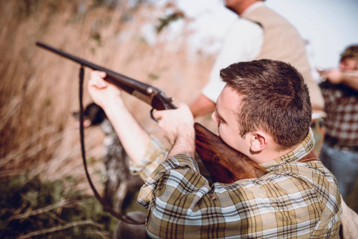 A man in a plaid shirt aims a shotgun outdoors with two others and a dog in the background, talking about when to buy Montana bonus points as a group.





