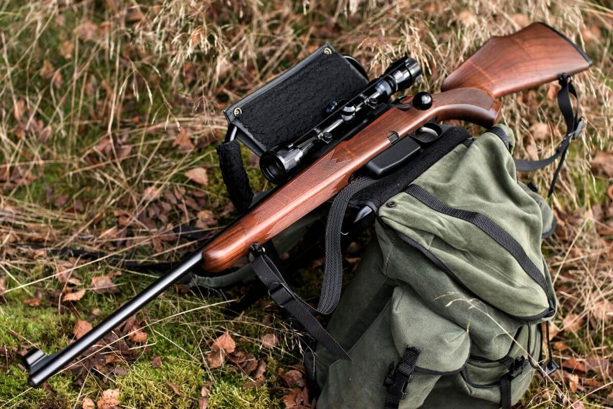 A hunting rifle with a scope attached, resting on a green canvas backpack on a grassy surface