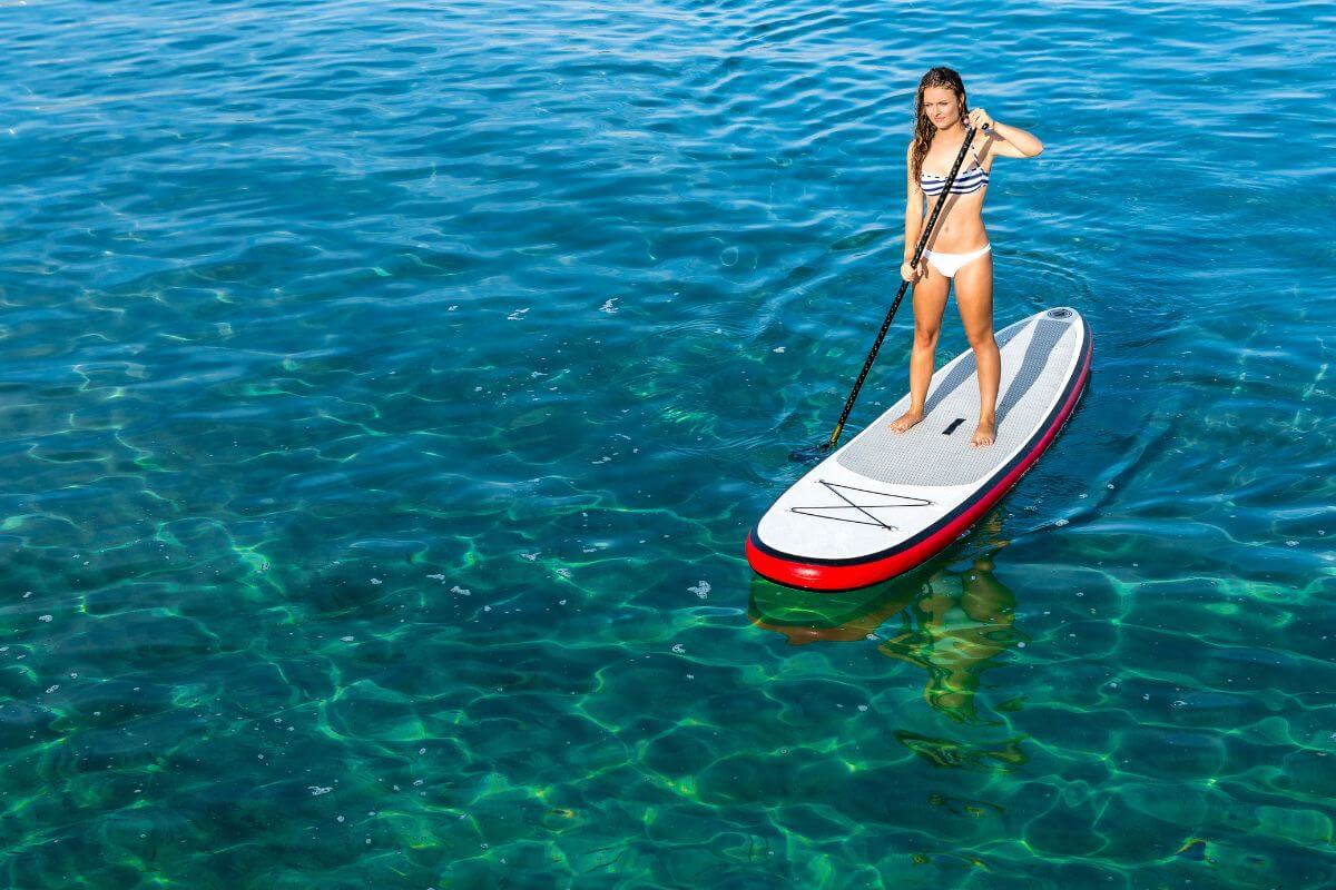 A woman in a striped bikini balances atop a white and red paddleboard in the clear blue waters near Monture Waterfall 