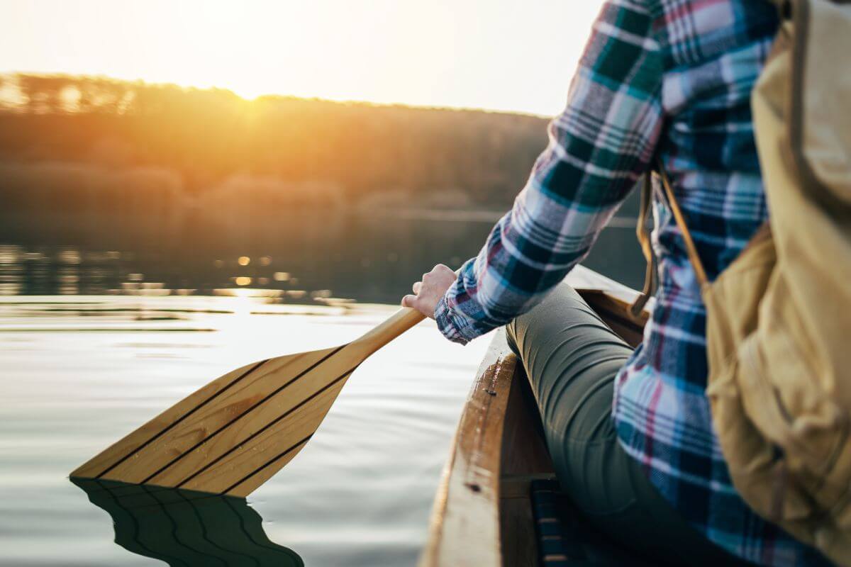 At sunset on a calm Montana lake, a person in a plaid shirt and beige backpack paddles a canoe. 